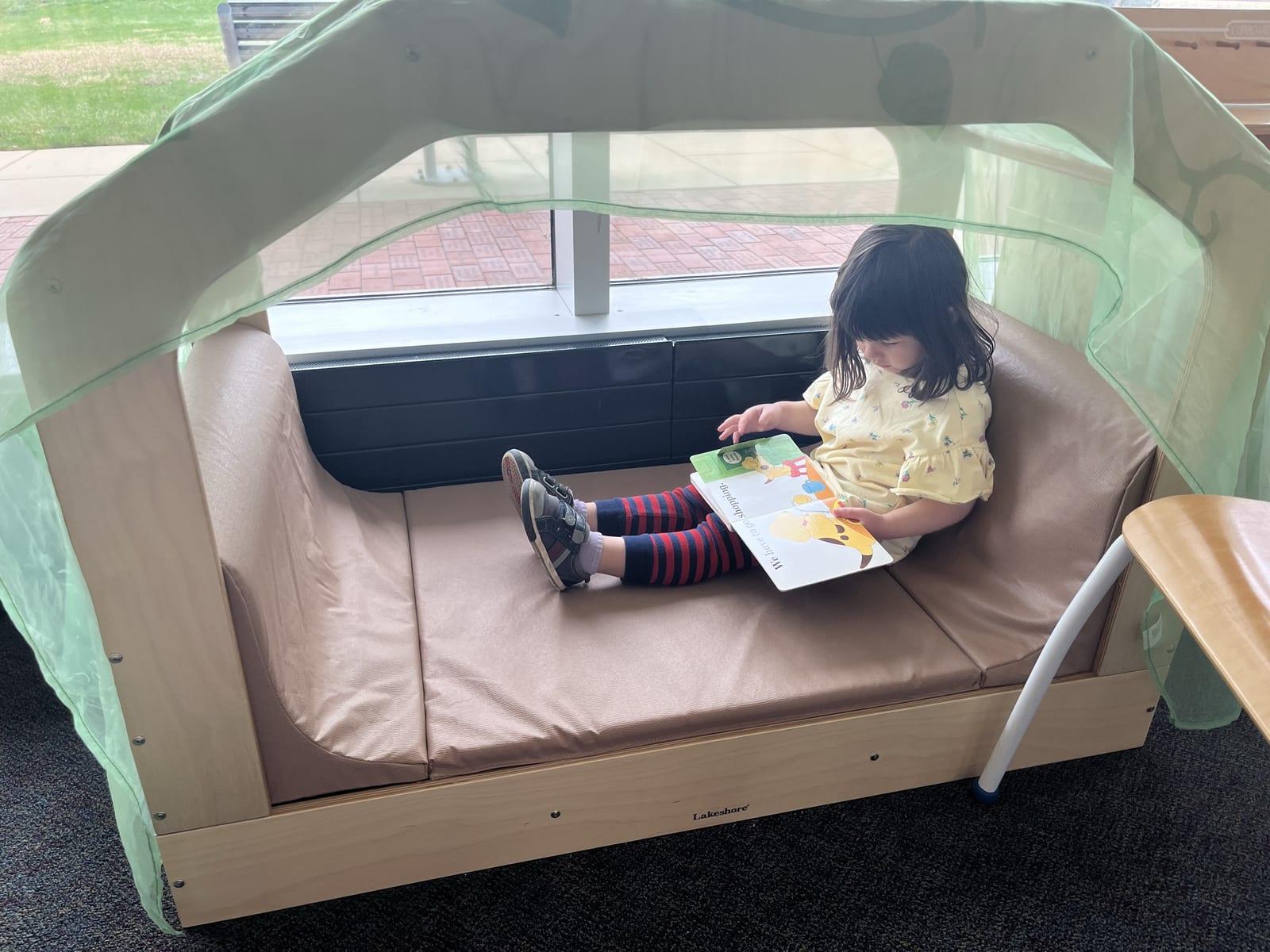 A child reading a picture book in a cushioned reading nook by the window at Brandywine Hundred Library
