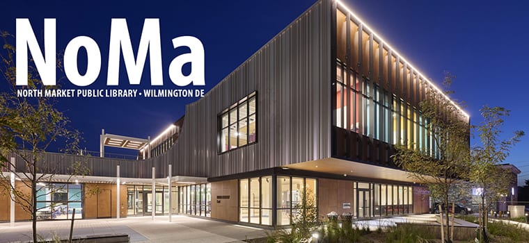 The new NoMa library at dusk — a two-story modern building on North Market Street in Wilmington, with warm-lit windows and colorful glass panels along the upper floor.