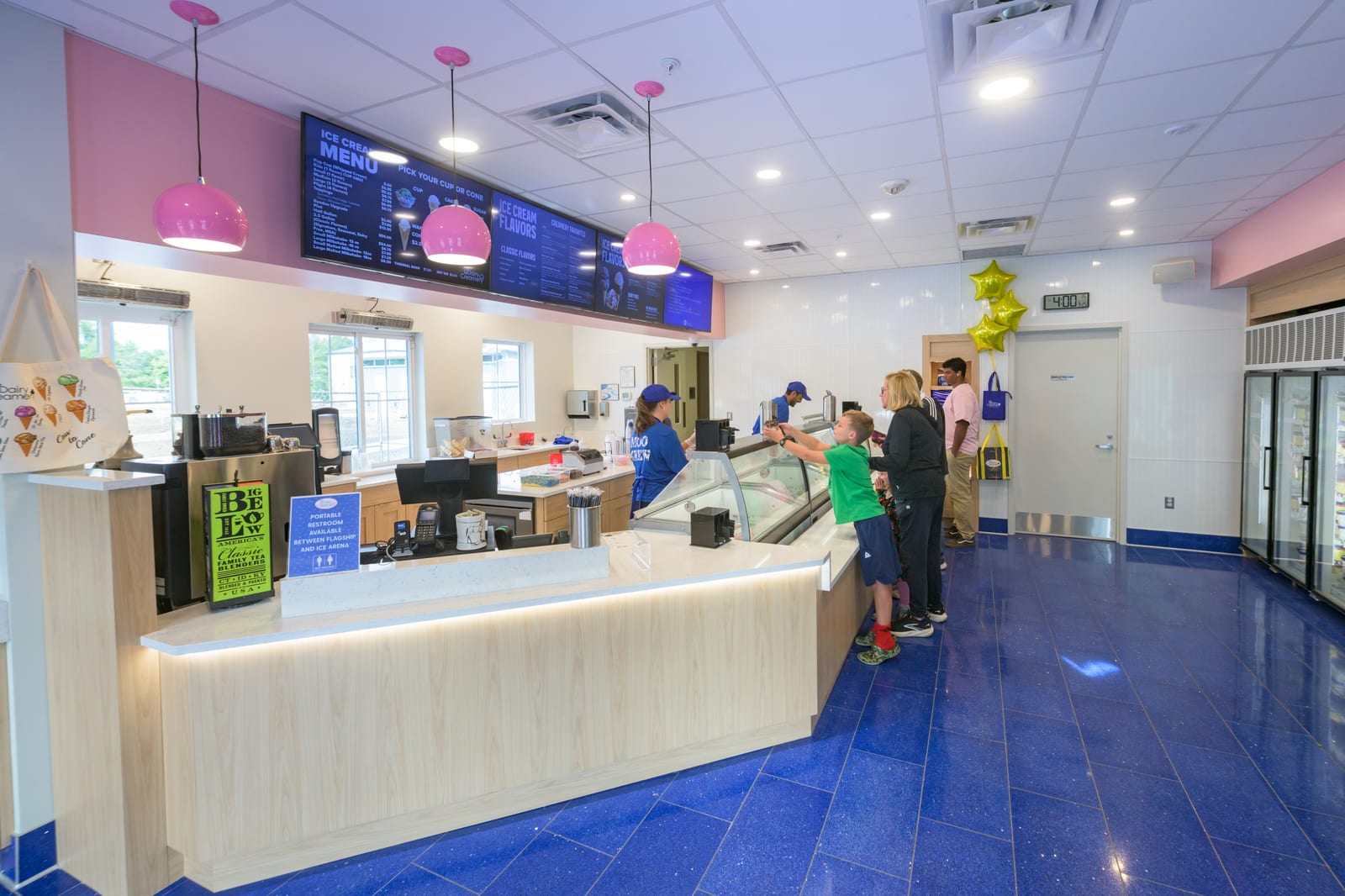 Inside the renovated UDairy Creamery: a boy in a green shirt points at the ice cream case while students in blue UDairy shirts scoop, with pink pendant lights and the menu boards overhead